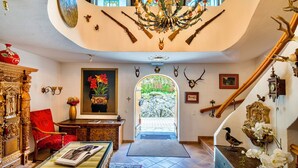 Un hall imponente con una araña, un sillón rojo, un aparador de madera y una escalera que sube a un nivel superior.