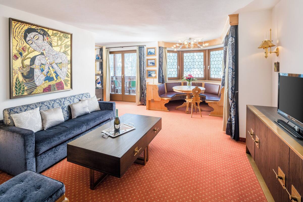 Una sala de estar con sofá, mesa de centro y televisor. Hay un cuadro en la pared y una ventana con cortinas.