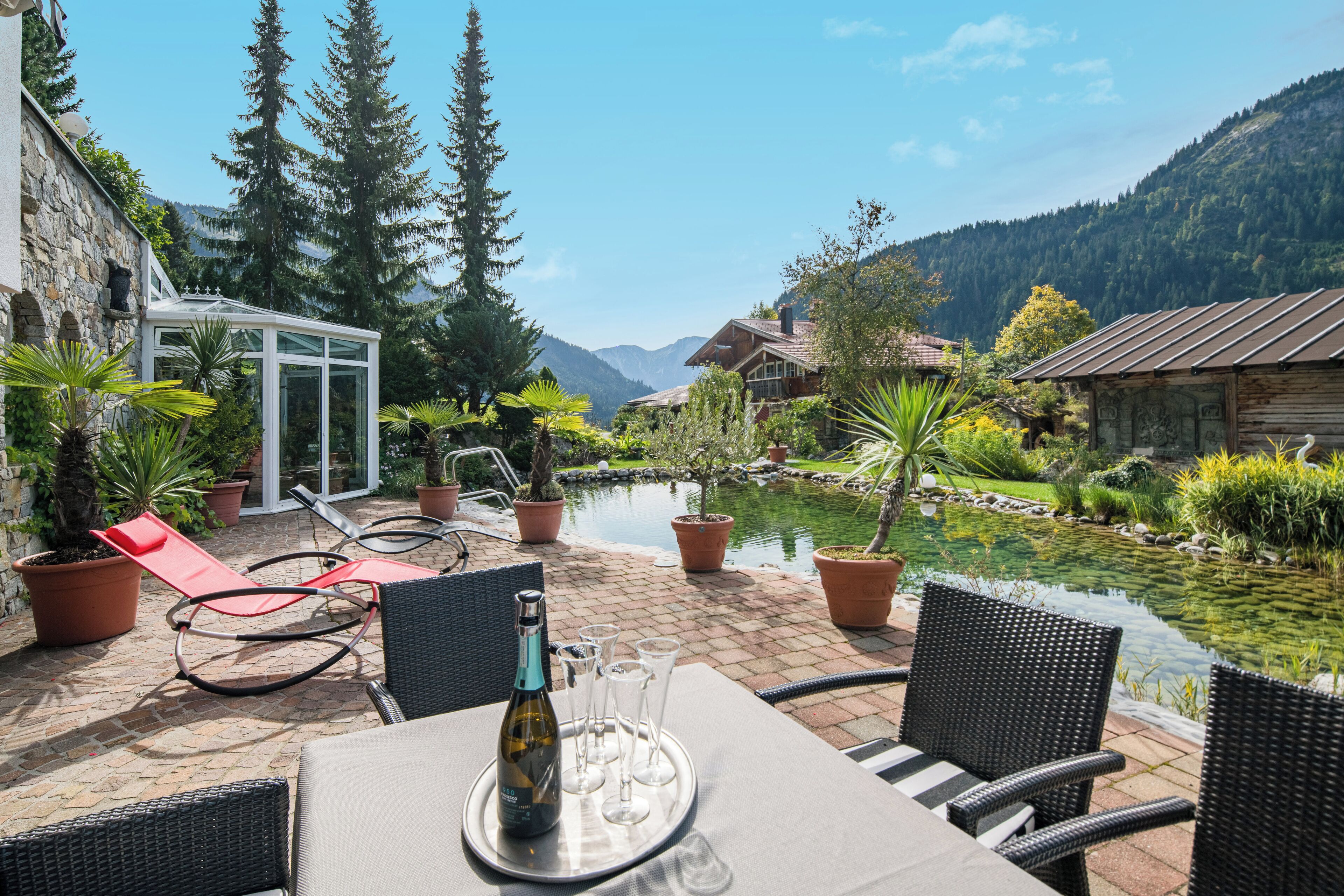 A patio with a table set for dining, surrounded by potted plants and chairs, overlooking a mountain and a pond.
