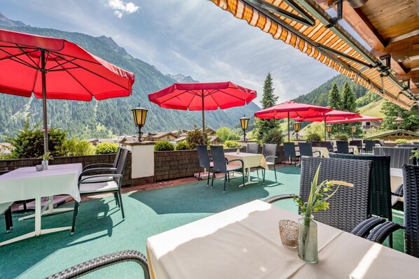 An outdoor patio with tables and chairs, red umbrellas, and a view of mountains.