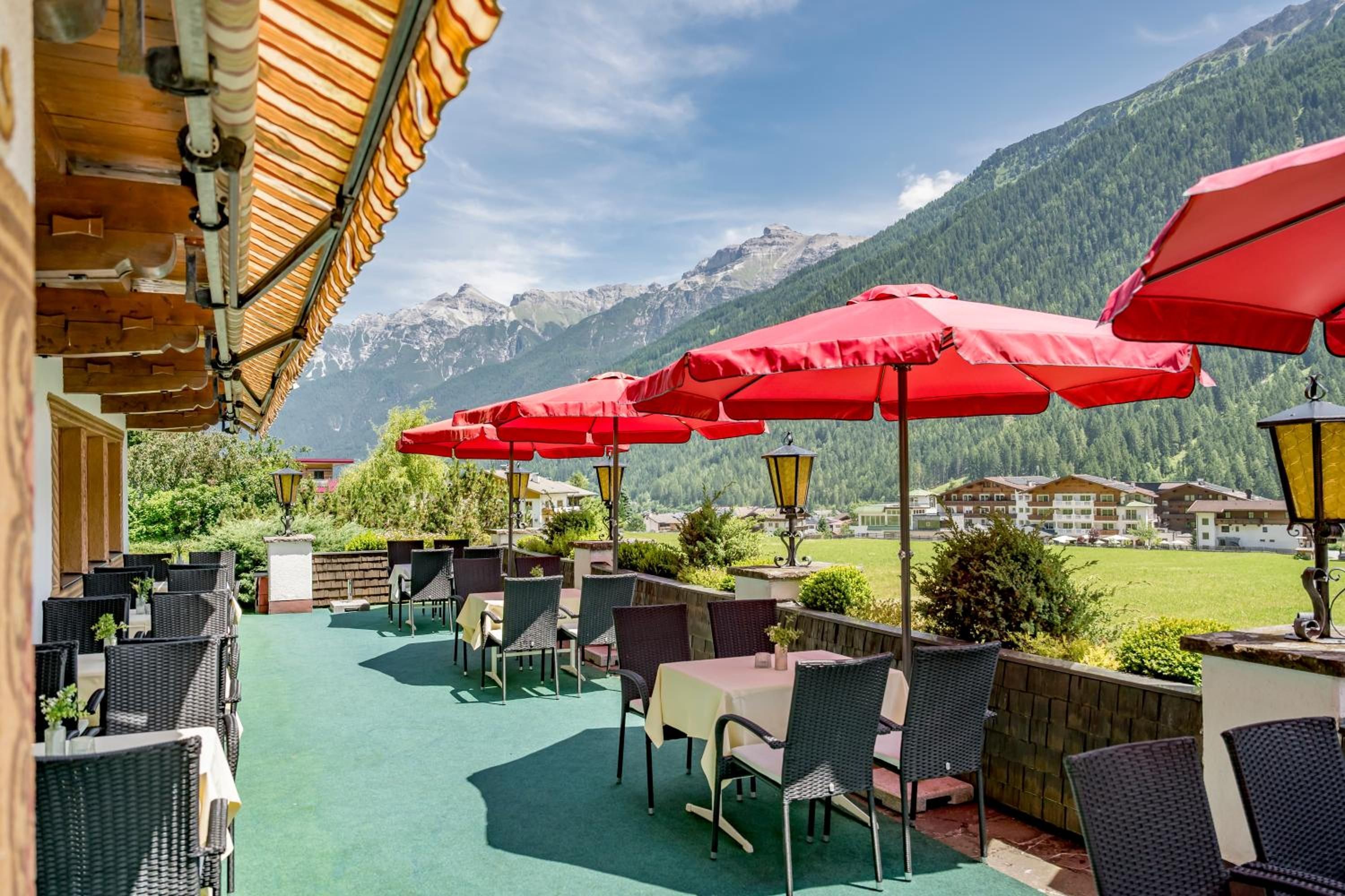 An outdoor seating area with tables and chairs, red umbrellas, and a view of mountains in the background.