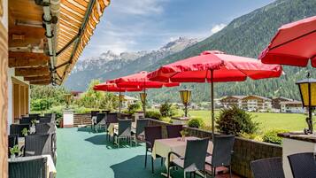 An outdoor seating area with tables and chairs, red umbrellas, and a view of mountains in the background.