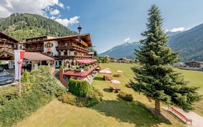 A traditional alpine hotel with a restaurant terrace, surrounded by lush greenery and mountains.