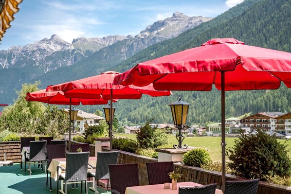 Outdoor seating with red umbrellas, wicker chairs, and tables, overlooking a mountainous landscape.