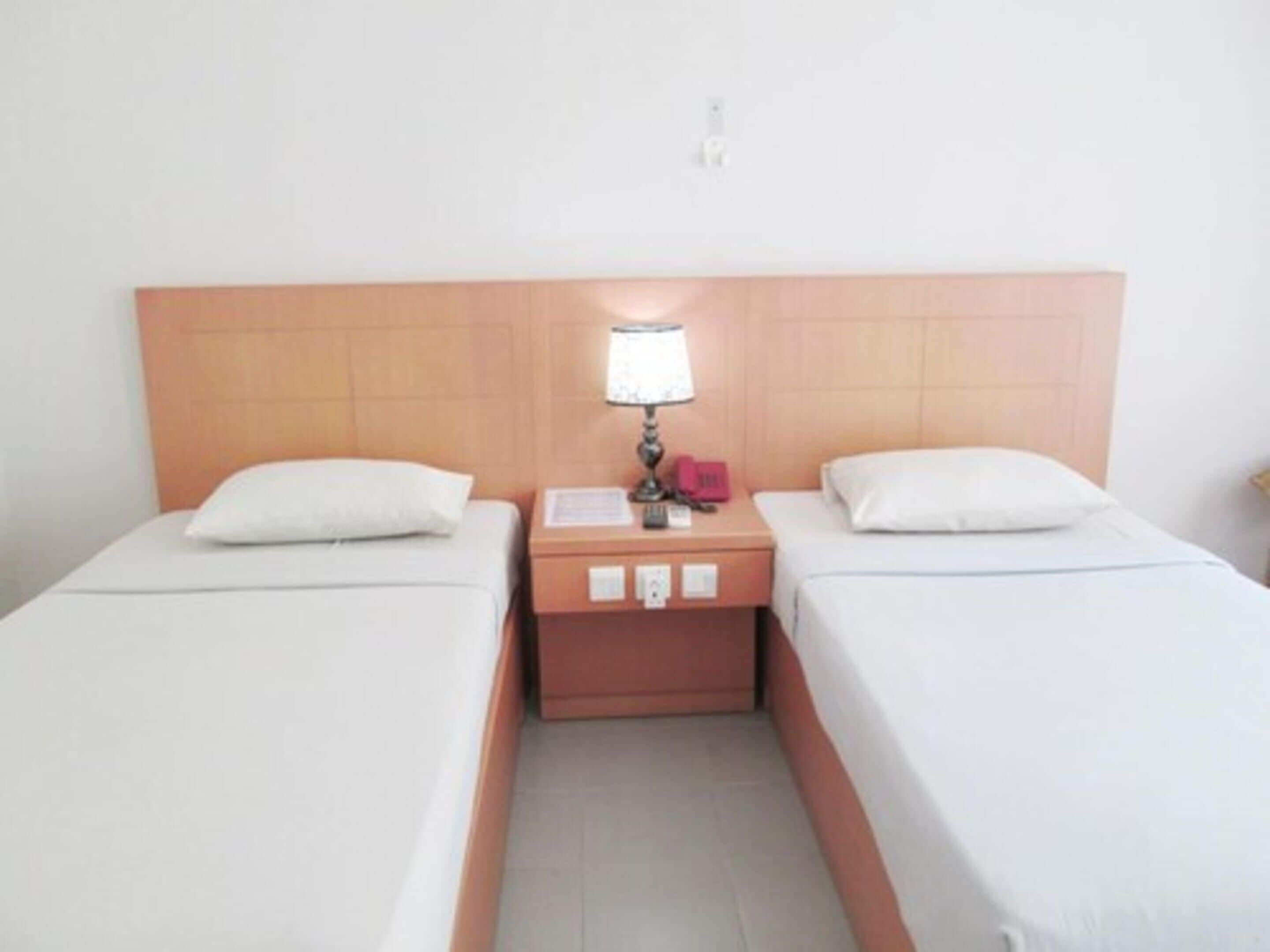 A hotel room with two single beds, a nightstand with a lamp, and a wall-mounted light fixture.