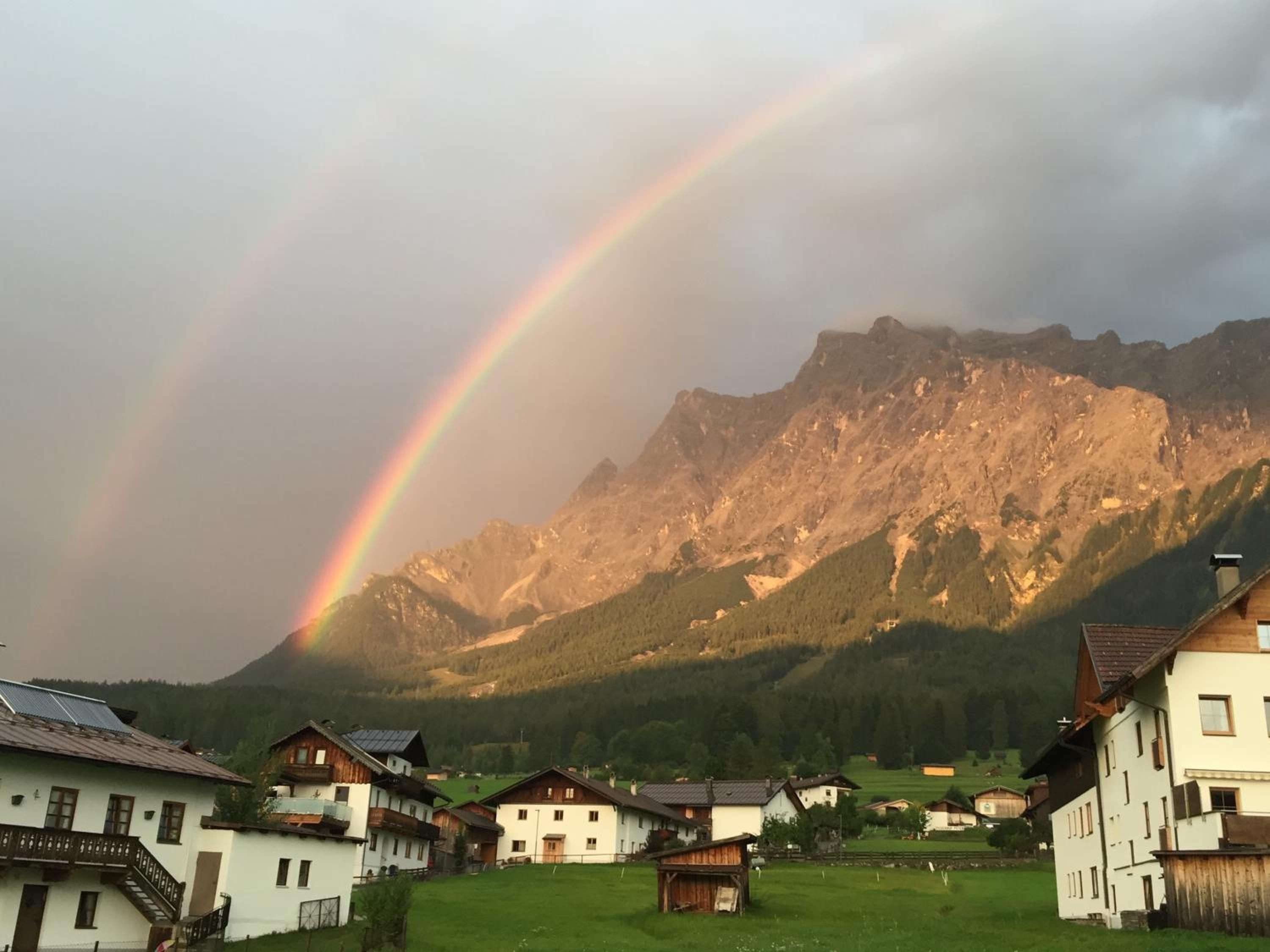 Ein ländliches Dorf mit traditionellen Häusern, Bergen im Hintergrund und einem deutlich sichtbaren Regenbogen.