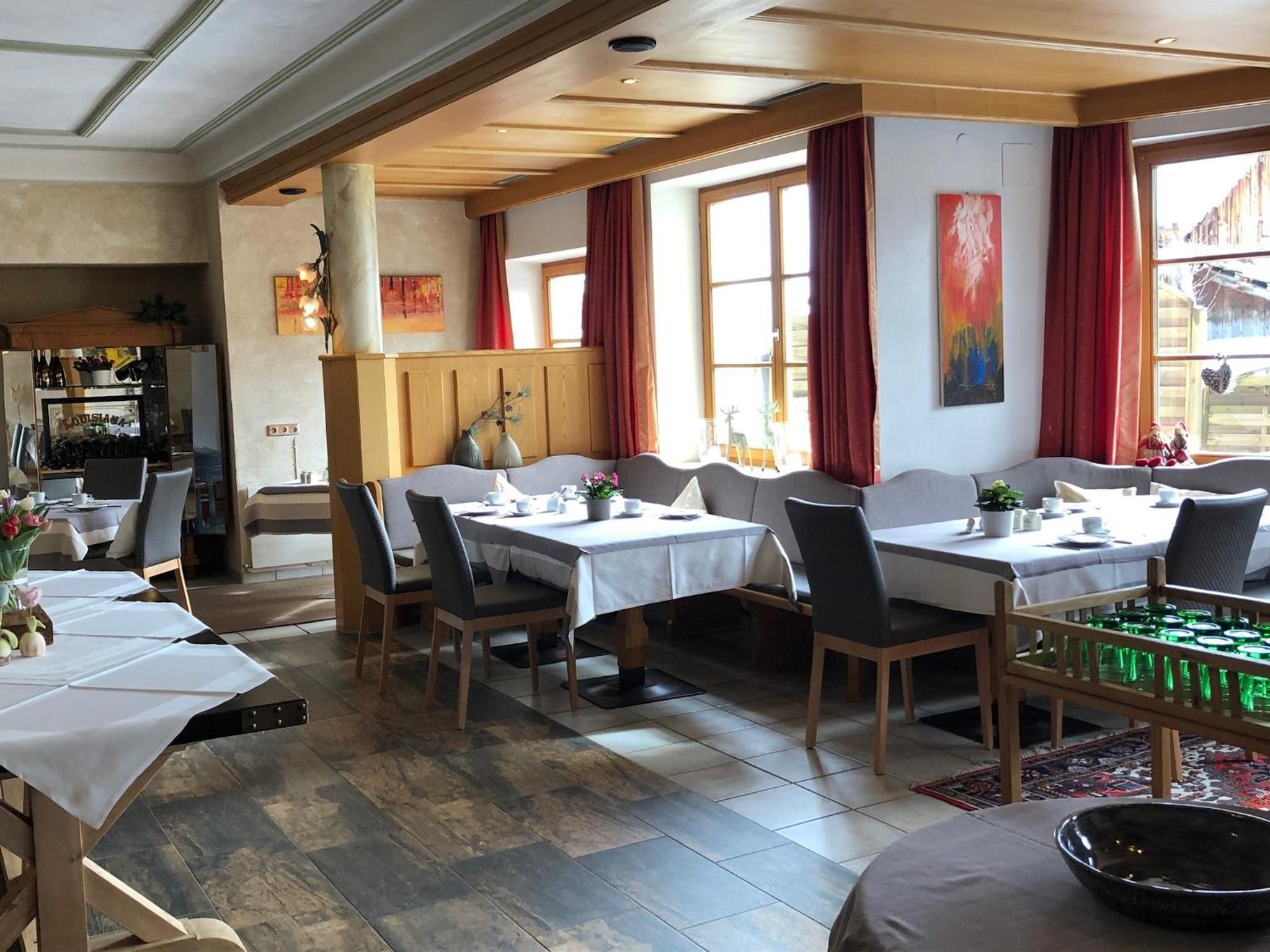 A dining area with multiple tables and chairs, wooden beams on the ceiling, and large windows with red curtains.