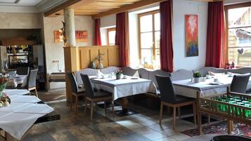 A dining area with multiple tables and chairs, wooden beams on the ceiling, and large windows with red curtains.
