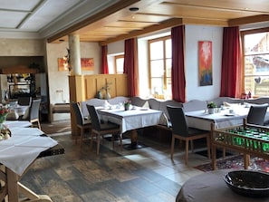A dining area with multiple tables and chairs, wooden beams on the ceiling, and large windows with red curtains. - Alpin (Ehrwald)