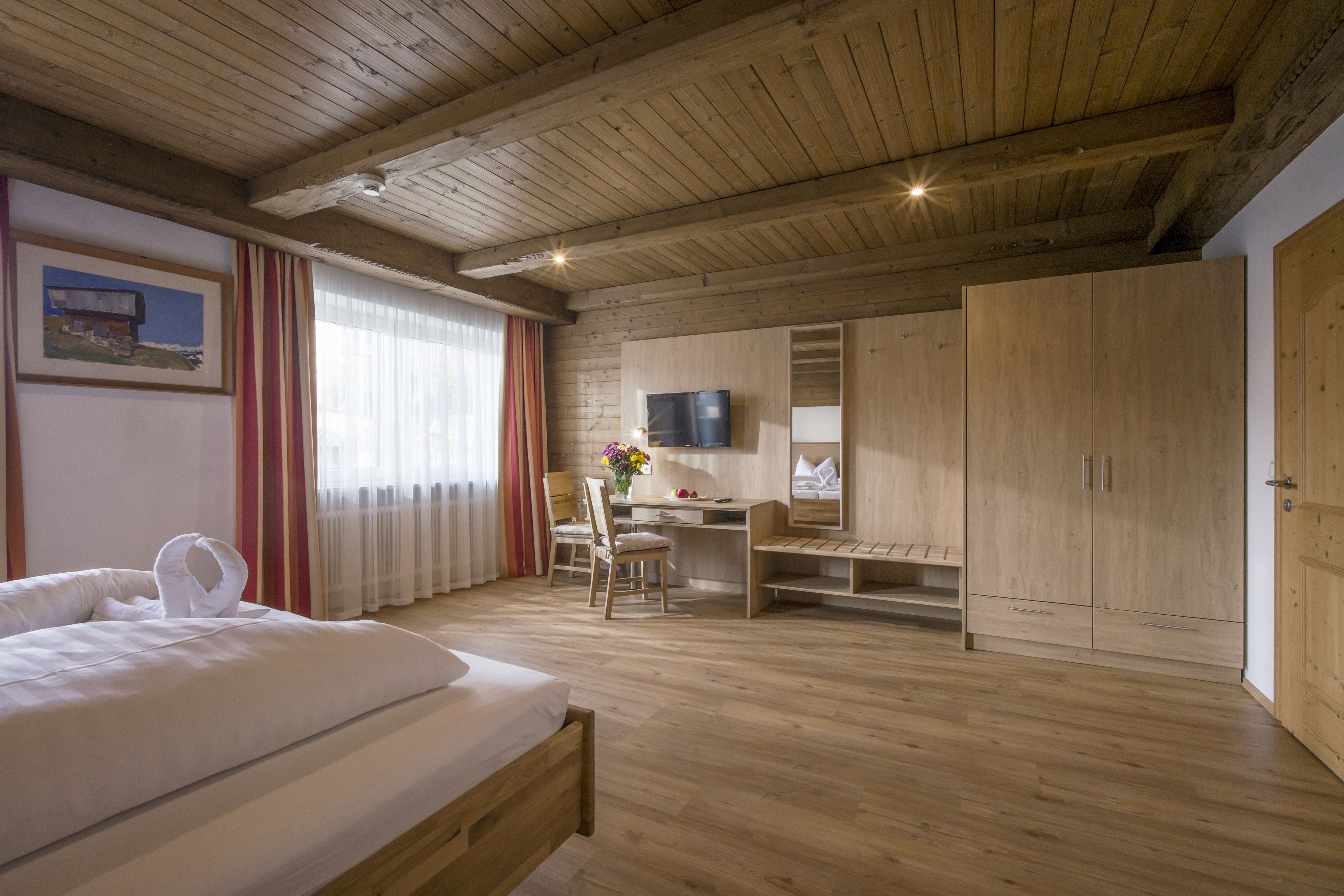 A hotel room with a wooden ceiling, a bed, a desk, a chair, a TV, and a wardrobe.