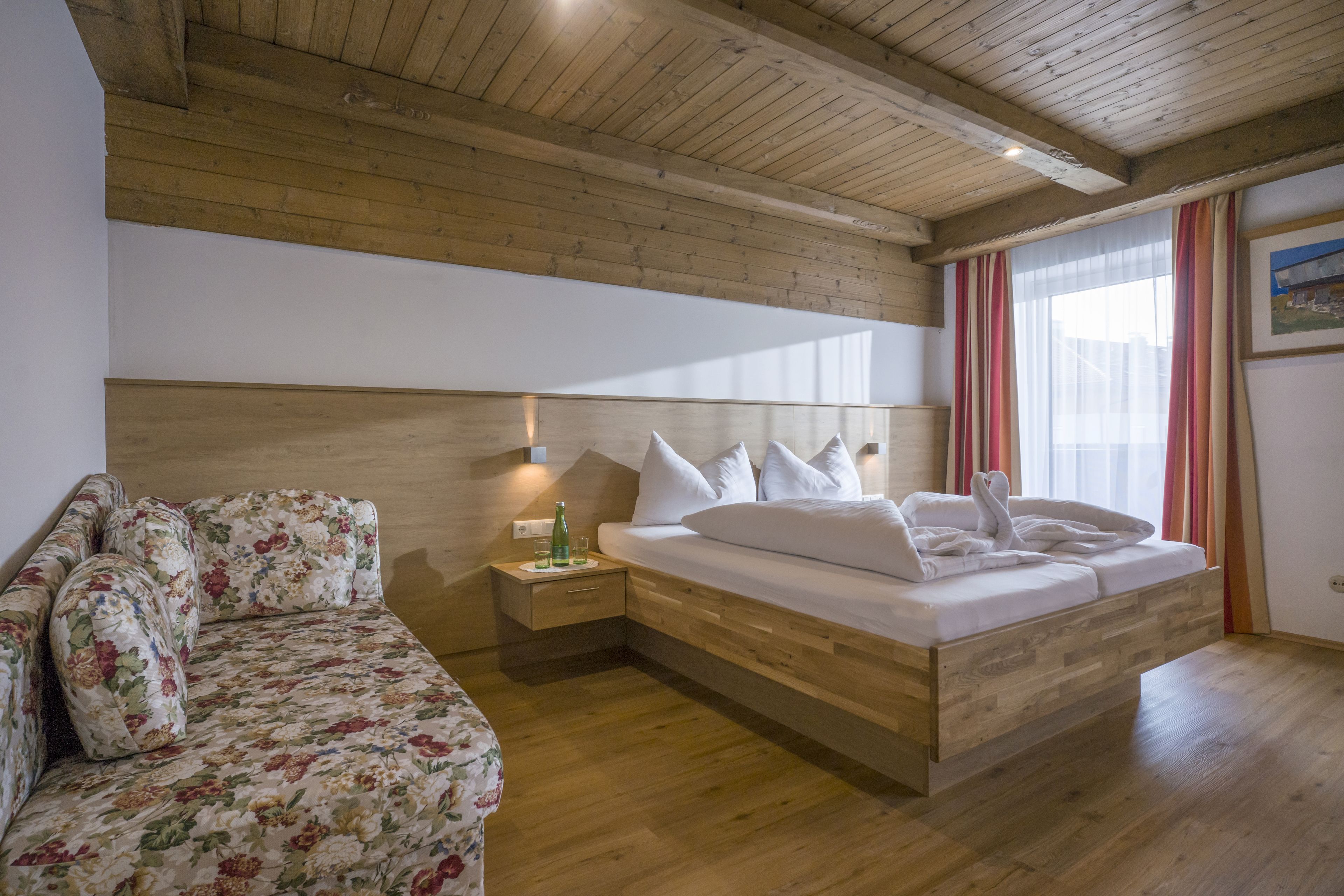 Een slaapkamer met een houten plafond, een bed met witte lakens, een bloemenbank en een nachtkastje met een fles.