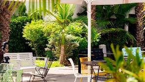 An outdoor seating area with a white umbrella, various chairs, and a palm tree. - Eden Inn (Zikhron Ya'aqov)