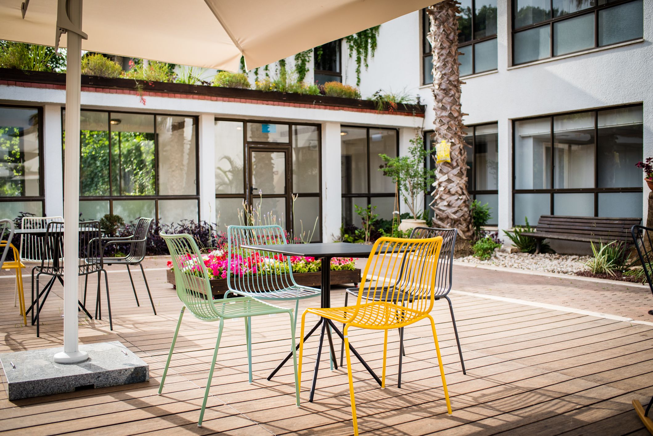 Un patio al aire libre con una mesa y sillas, rodeado de plantas y un edificio con ventanas grandes.