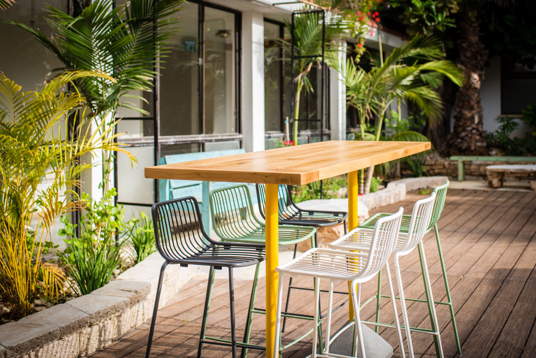Un patio al aire libre con una mesa de madera y sillas de metal, rodeado de follaje y un edificio con grandes ventanales.