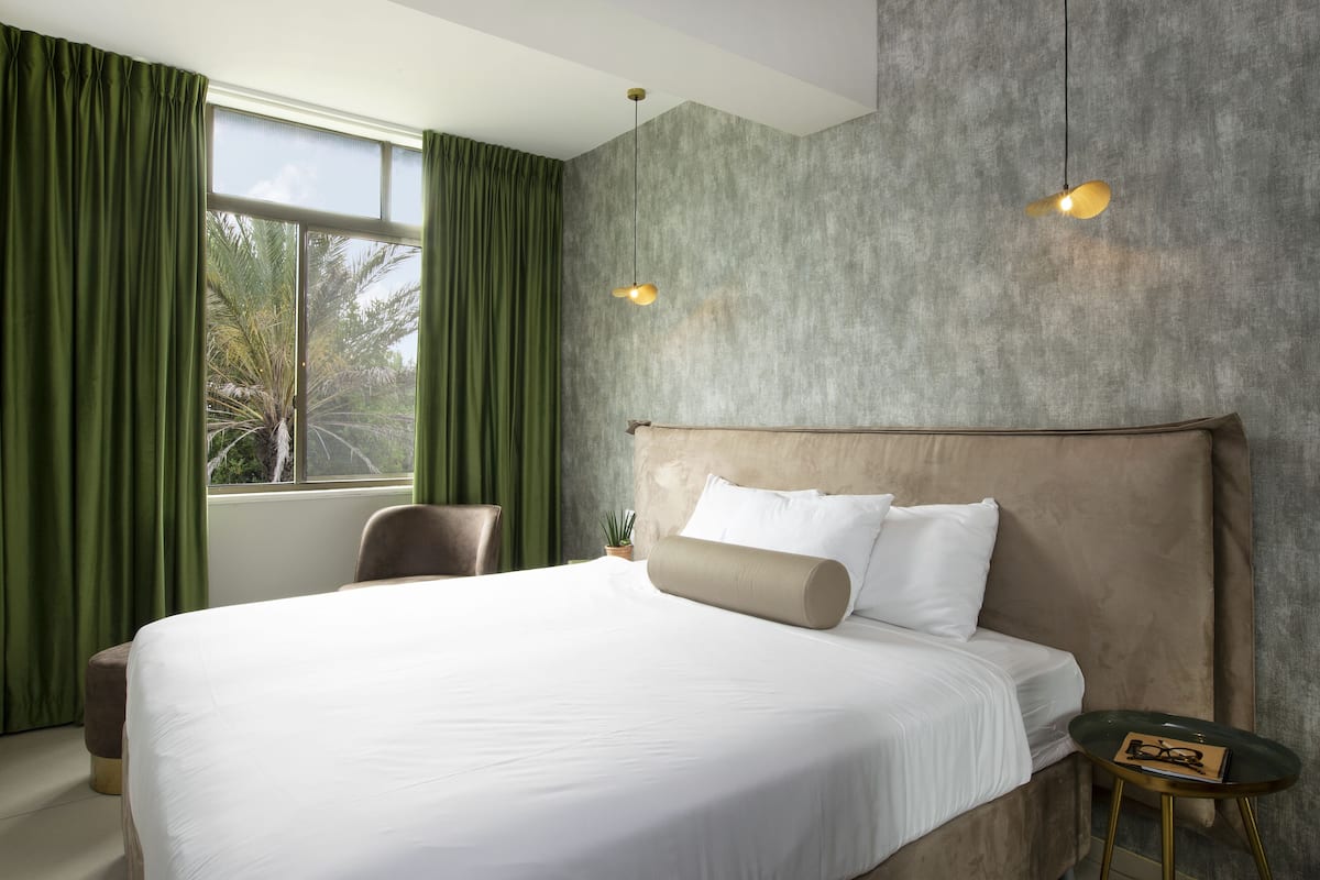 A hotel room with a large bed, green curtains, a headboard, a small table, and a hanging light fixture.