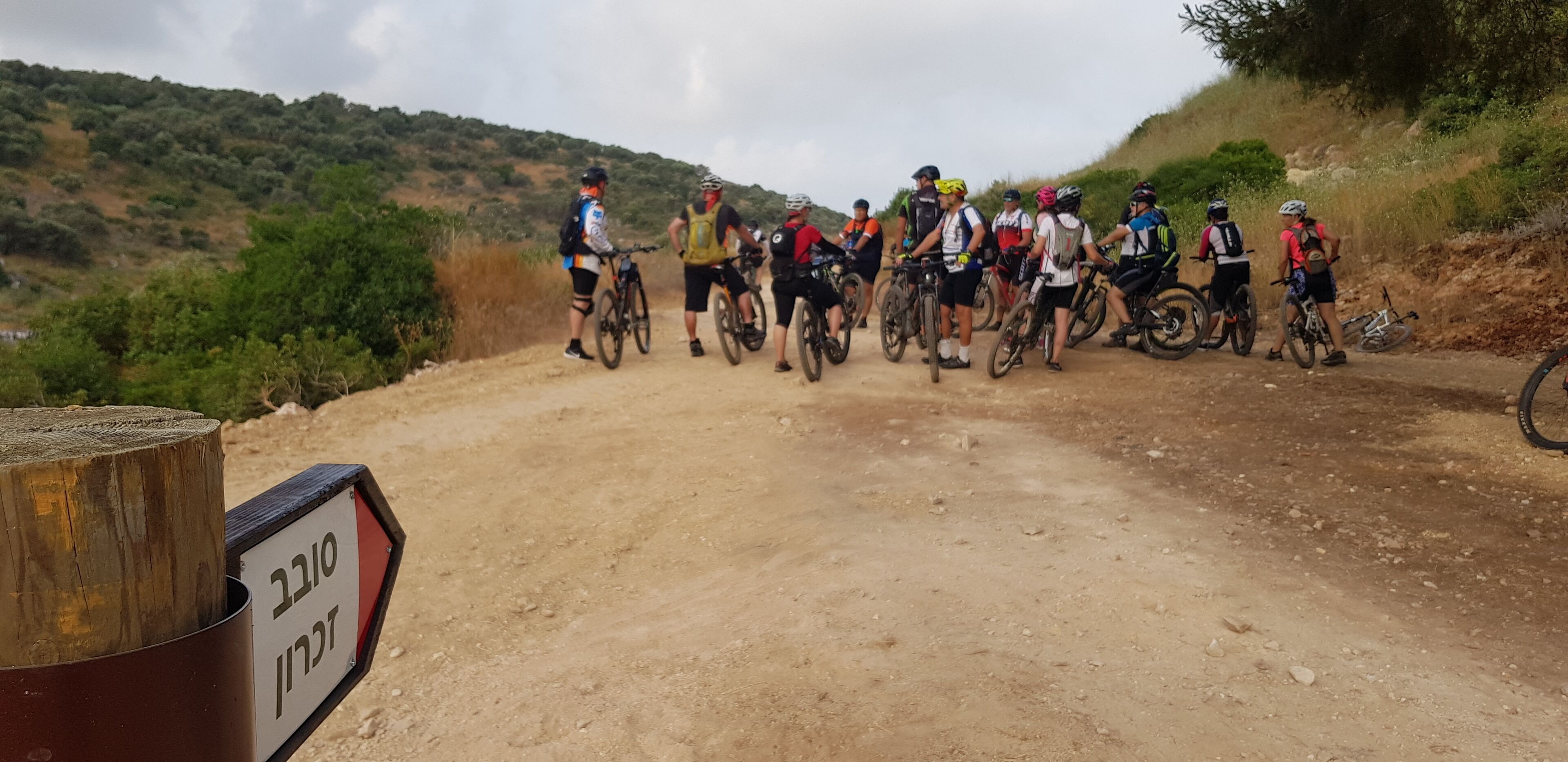 A group of cyclists on a dirt path with a wooden post and a sign in the foreground.