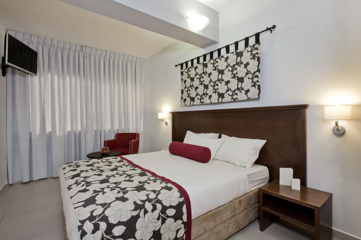 A hotel room with a large bed, a red chair, a small table, and a wall-mounted TV.