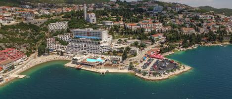 An aerial view of a coastal town with a prominent hotel featuring a swimming pool, a parking area, and a beach.