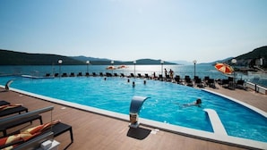 Une piscine entourée d’une terrasse en bois, agrémentée de chaises longues et de parasols, et offrant une vue sur la mer et les collines.
