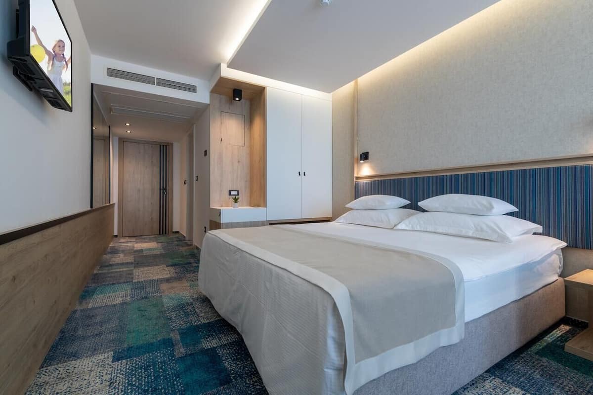 A modern hotel room with a large bed, a flat-screen TV mounted on the wall, a bedside table with a lamp, and a patterned carpet.