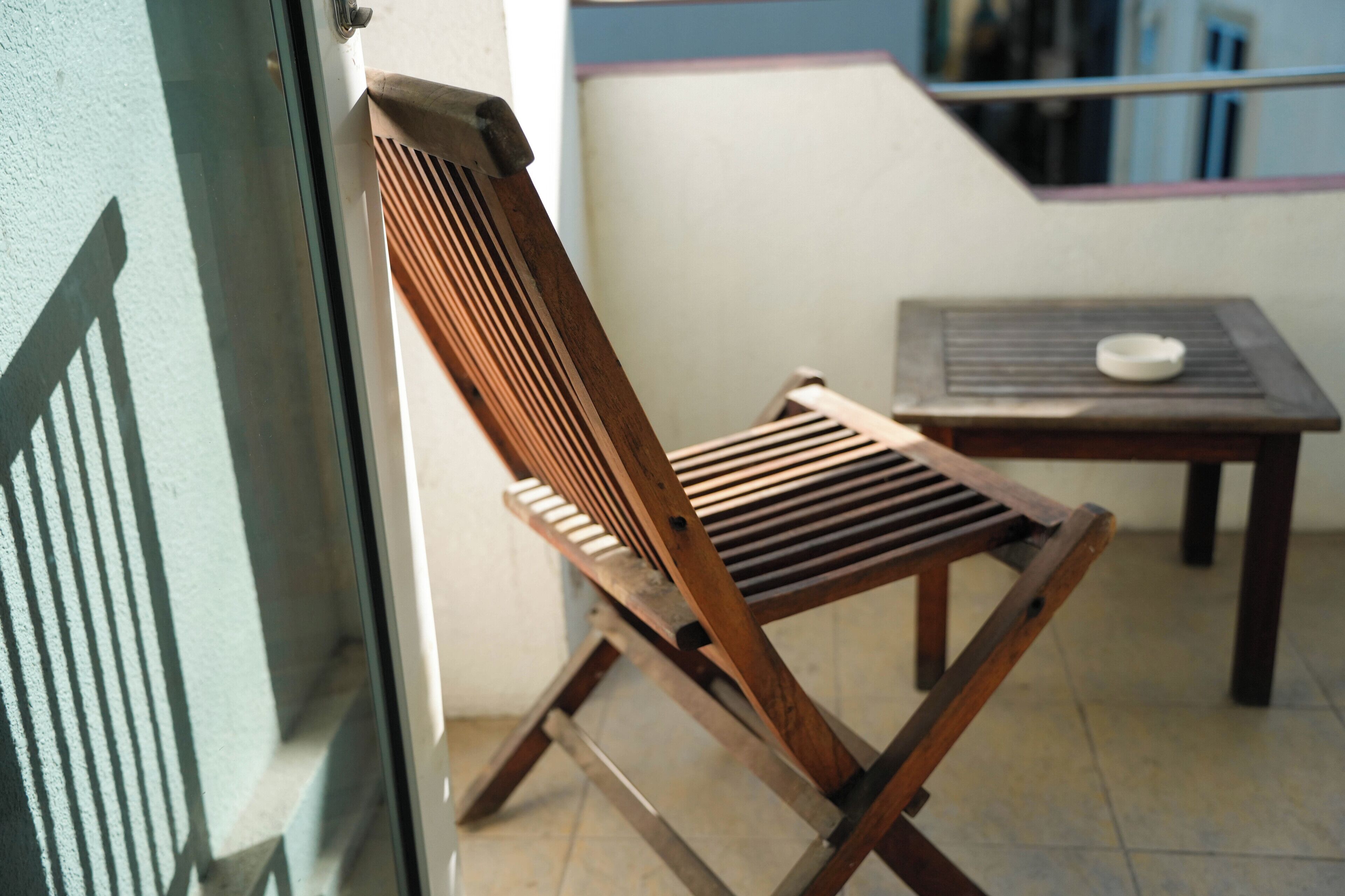 Una silla de madera con respaldo de listones y una pequeña mesa con un cuenco blanco, ubicadas en un balcón con vista a un edificio.