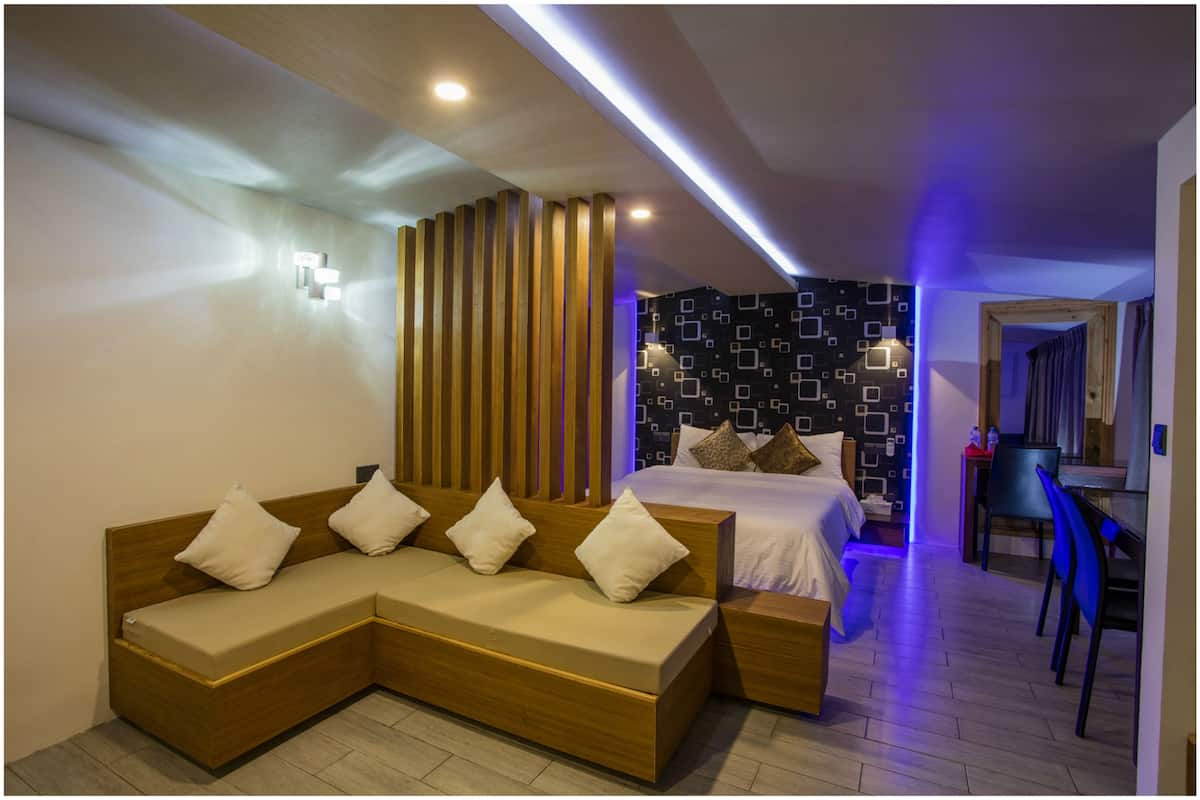 A modern hotel room with a wooden sectional sofa, a bed with a patterned headboard, and a dining area with a chair and table.
