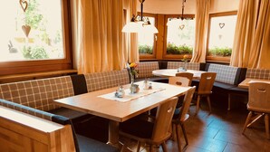 Een restaurant met houten meubels, waaronder zitjes en tafels, en grote ramen met gordijnen.