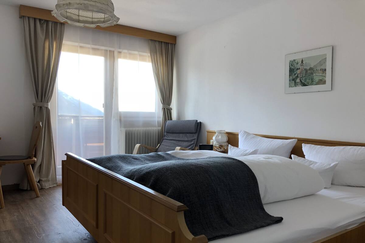 Une chambre d’hôtel avec un lit en bois, une chaise, une table de chevet avec une lampe et une fenêtre avec des rideaux.