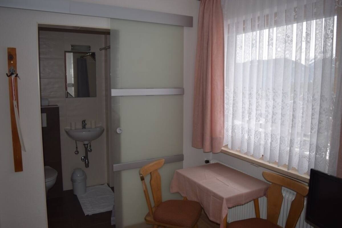 Une salle de bain avec un lavabo, un miroir et des toilettes. On y trouve une table recouverte d’une nappe et deux chaises.