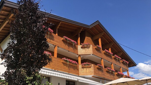 Een houten chalet met balkons versierd met bloemen, een terras met buitenmeubels, en een helderblauwe lucht.