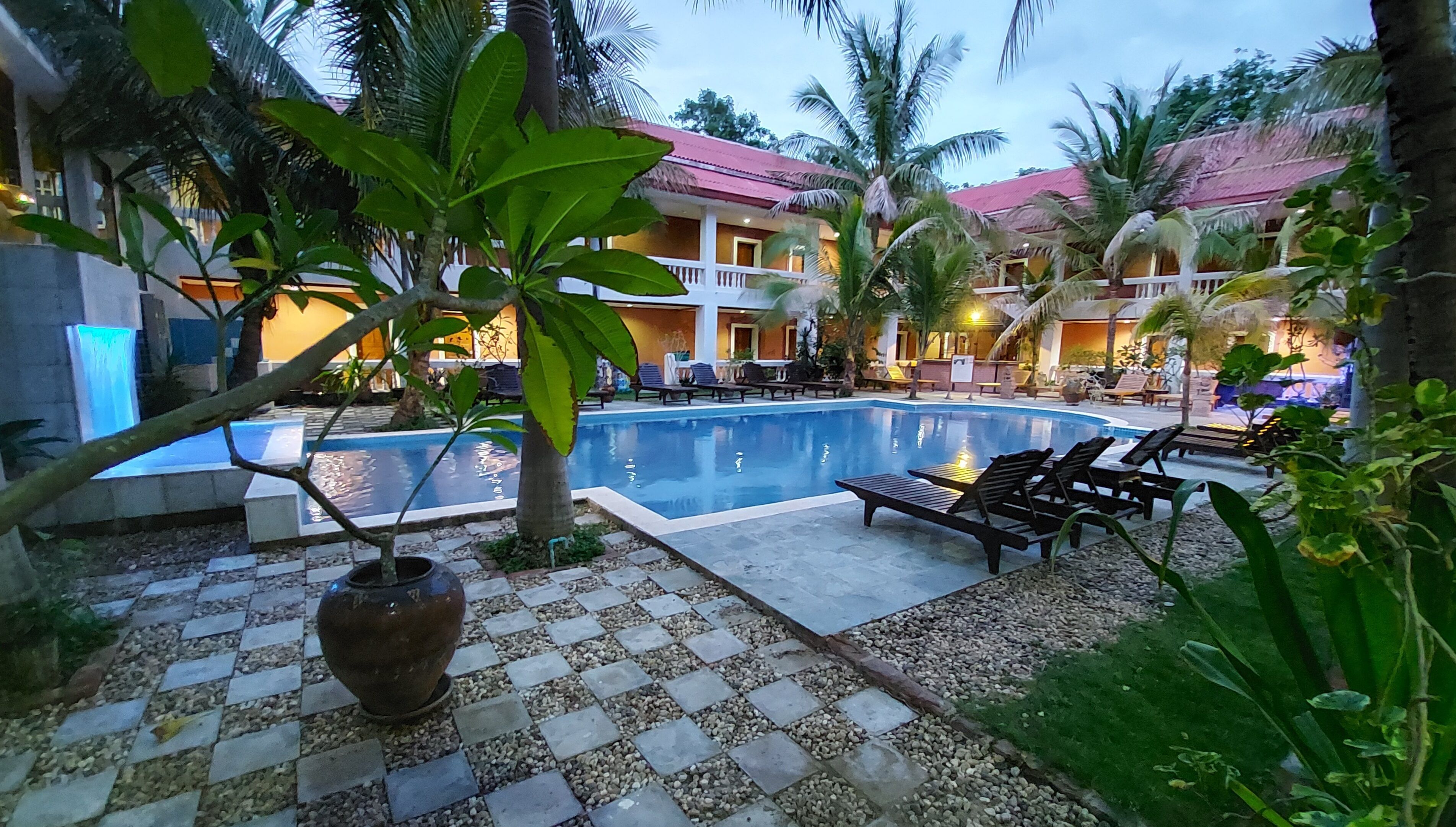 A swimming pool with lounge chairs, surrounded by palm trees and a building with a red roof.