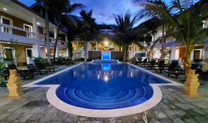 Una piscina con agua azul, rodeada de sillas de descanso y palmeras, con un edificio al fondo.
