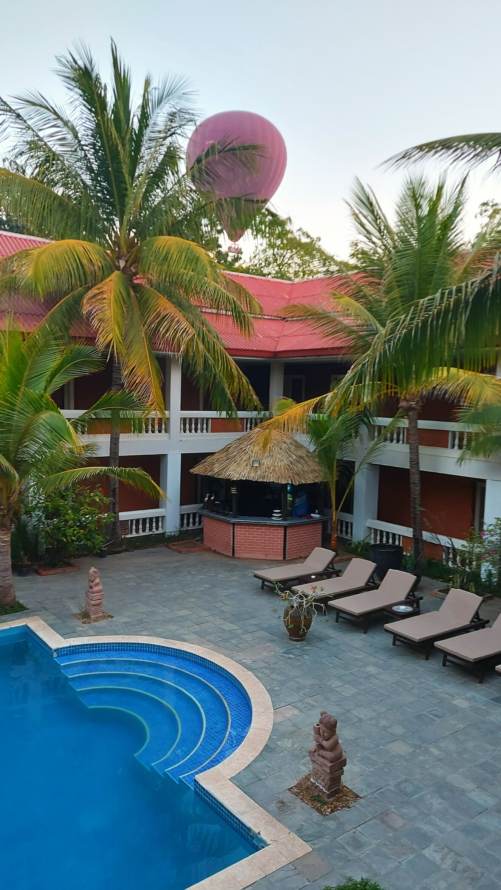 Ein Resort mit Swimmingpool, Liegestühlen, Palmen und einer Struktur mit einem Reetdach.