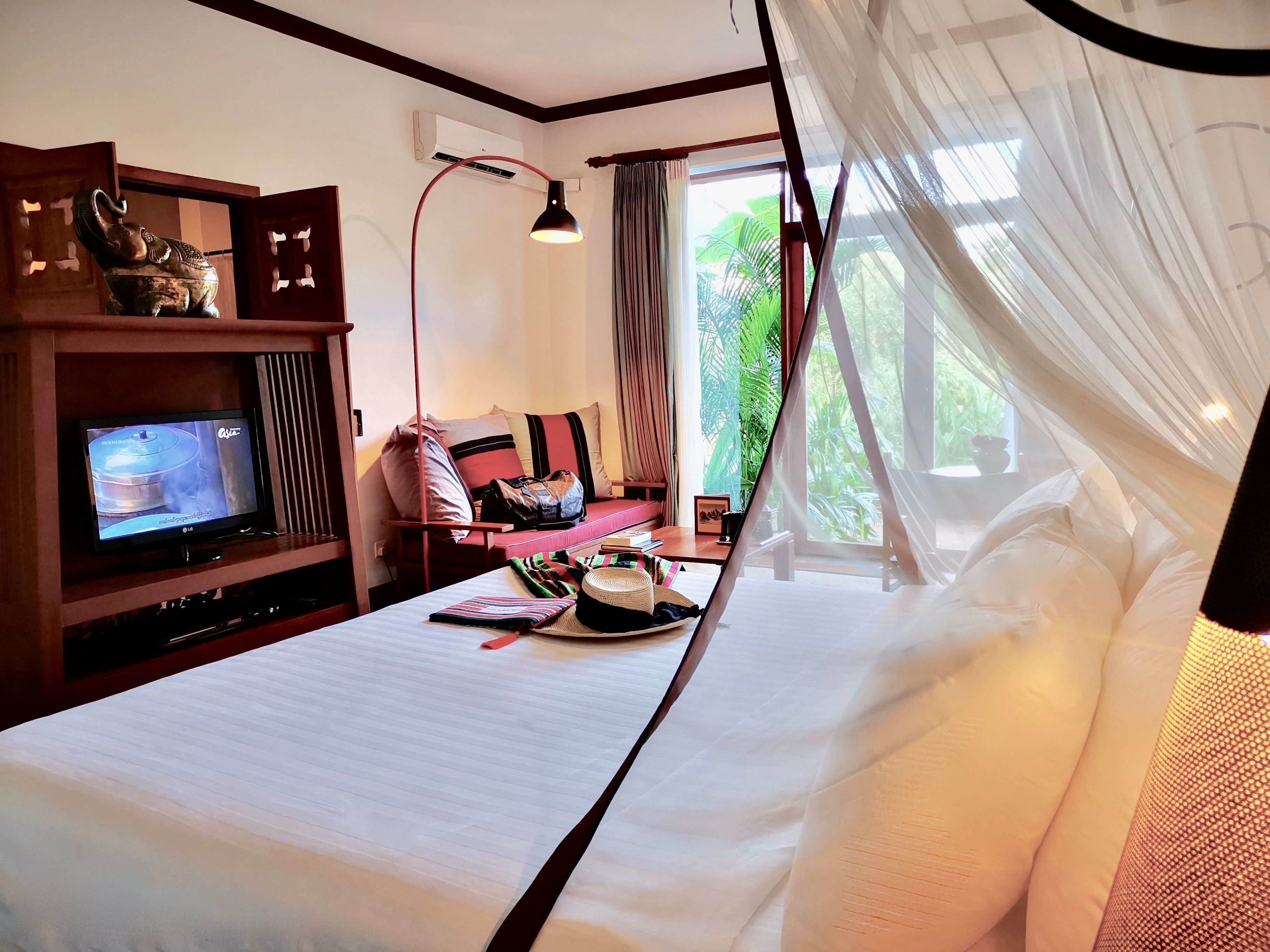 A hotel room with a bed, a television, a desk with a lamp, and a view of greenery outside.
