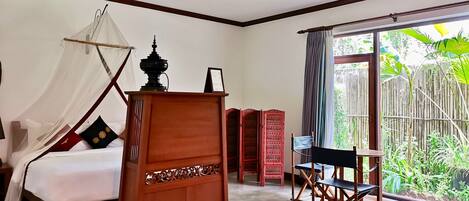 A bedroom with a bed, a wooden cabinet, a desk with a chair, and a large window with a view of greenery.