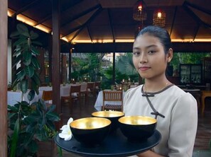 A person holding a tray with three bowls, in a restaurant with wooden tables and chairs, and a covered outdoor area with plants. - Blue Bird (Nyaung-U)