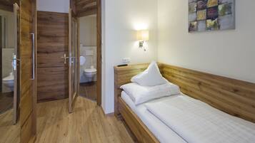 Une chambre d’hôtel avec un plancher en bois, un lit avec une literie blanche, une tête de lit en bois, une lampe fixée au mur et une porte en bois donnant accès à la salle de bain.