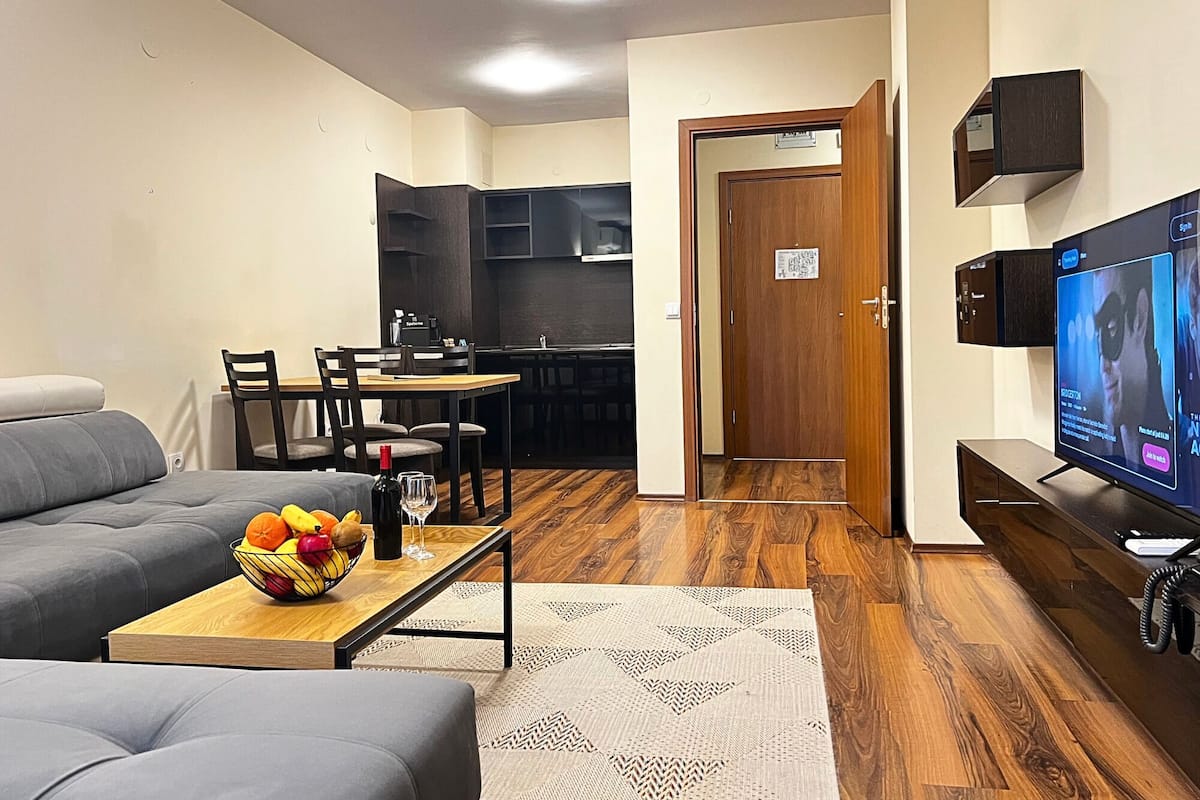A living room with a sofa, a dining table with chairs, a TV, and a kitchen area with cabinets.