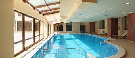 An indoor swimming pool with a glass ceiling, sunlit interior, and lounge chairs along the poolside.
