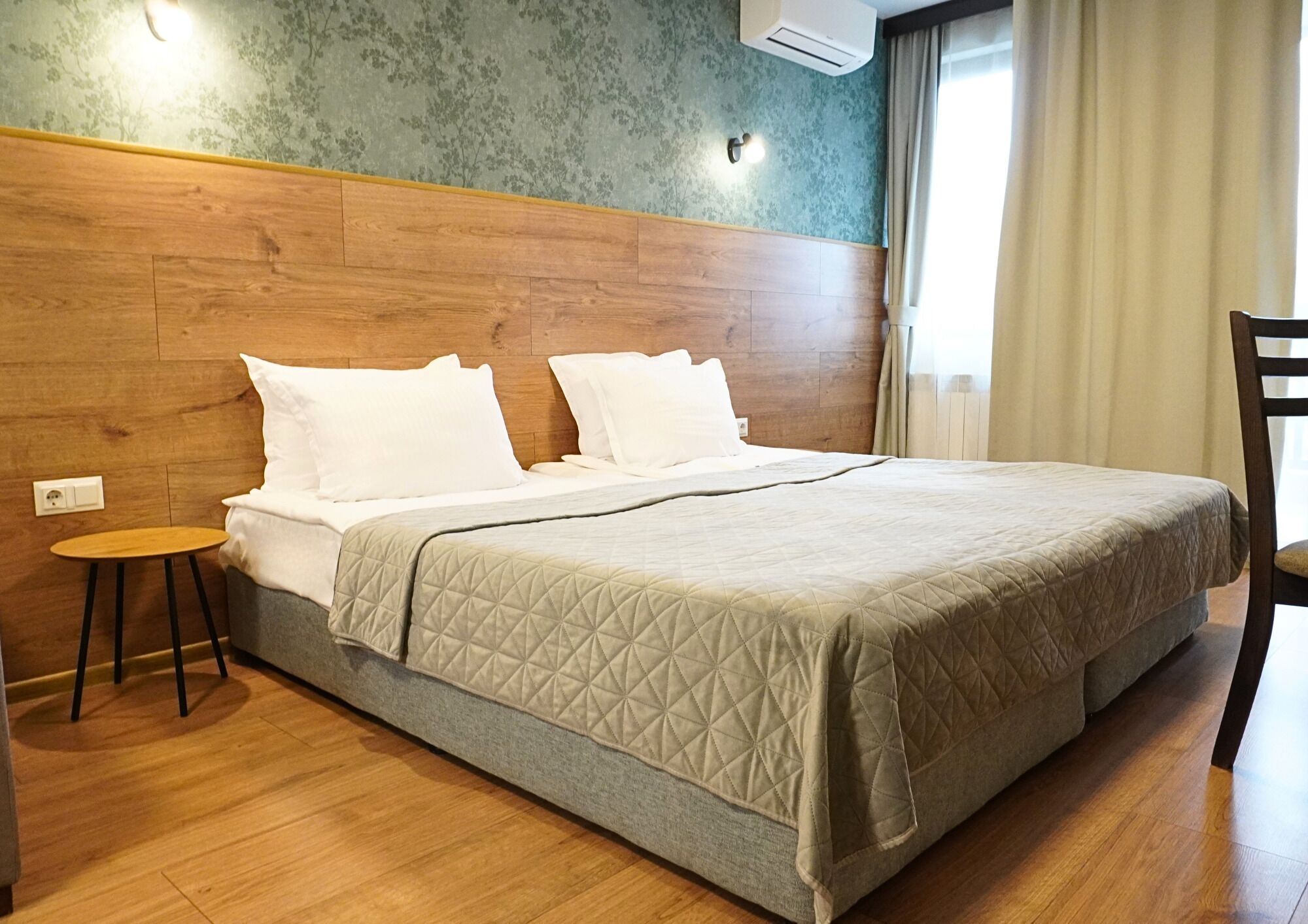 Una habitación de hotel con cabecera de madera, cama con colcha acolchada, una mesita redonda y una silla.