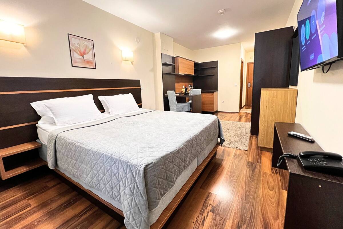 A hotel room with a bed, bedside table, TV, and a desk with a phone.