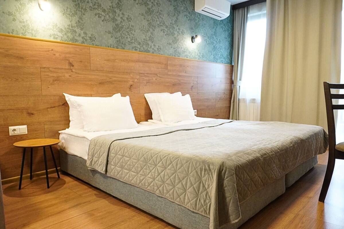 A hotel room with a wooden headboard, a bed with a quilted bedspread, a small round table, and a chair.