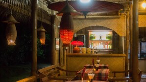 Un área de comedor al aire libre con mobiliario y mesas de madera, una barra con botellas y faroles colgantes.