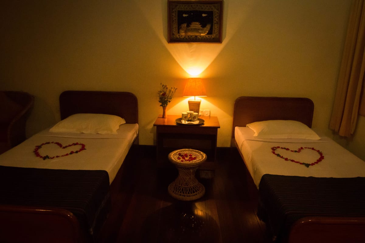 A hotel room with two single beds, each with a heart-shaped decoration on the bedspread. There is a small table with a vase of flowers between the beds. A wall-mounted lamp provides light, and a framed picture hangs above the table.