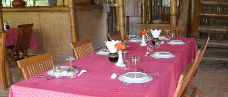 A dining area with a long table set for four, covered with a pink tablecloth. There are wooden chairs around the table, and a bamboo structure in the background.