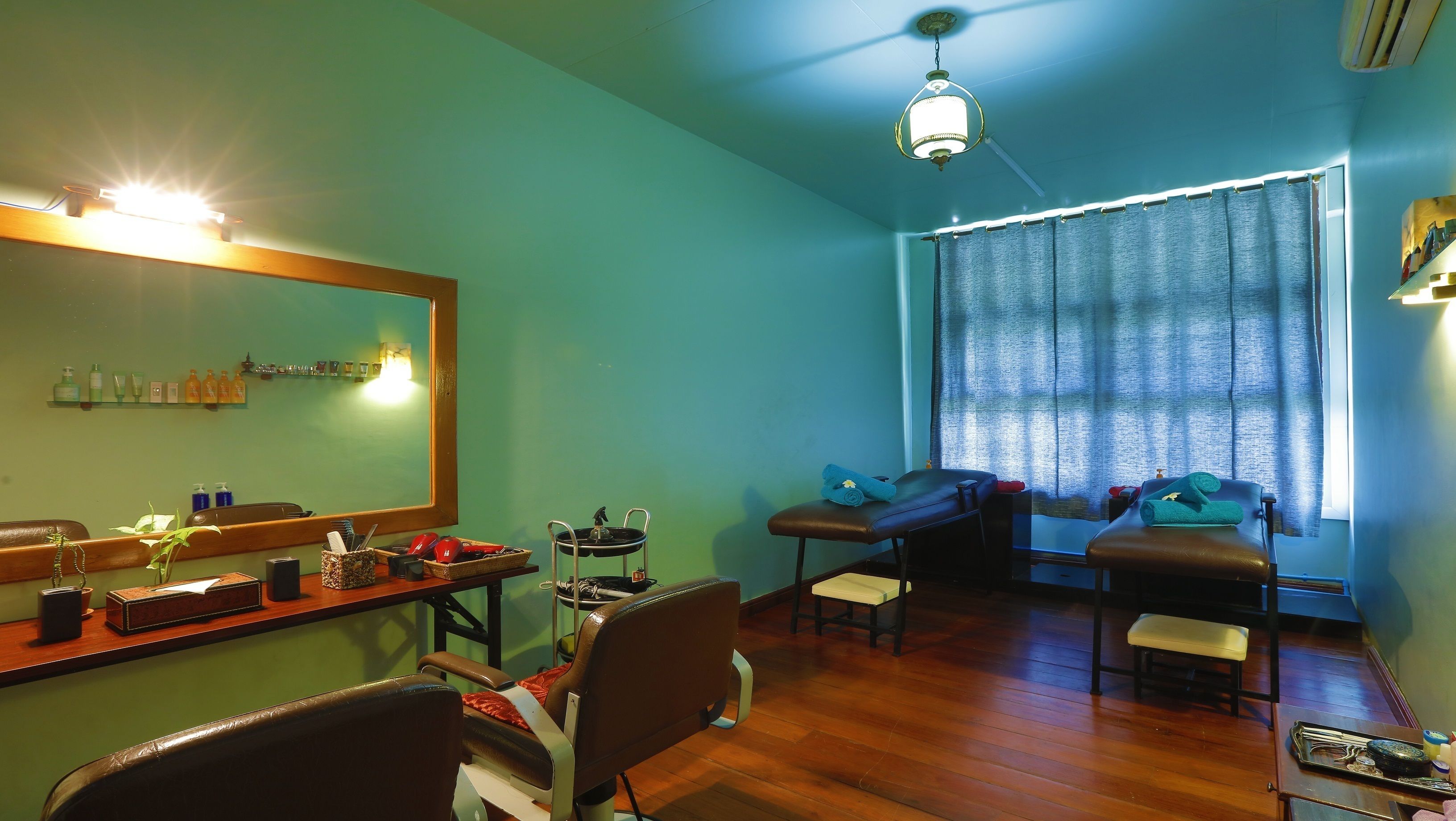 A room with a wooden floor, a large mirror, a vanity with toiletries, two massage chairs, a small table with a tray, and a window with curtains.