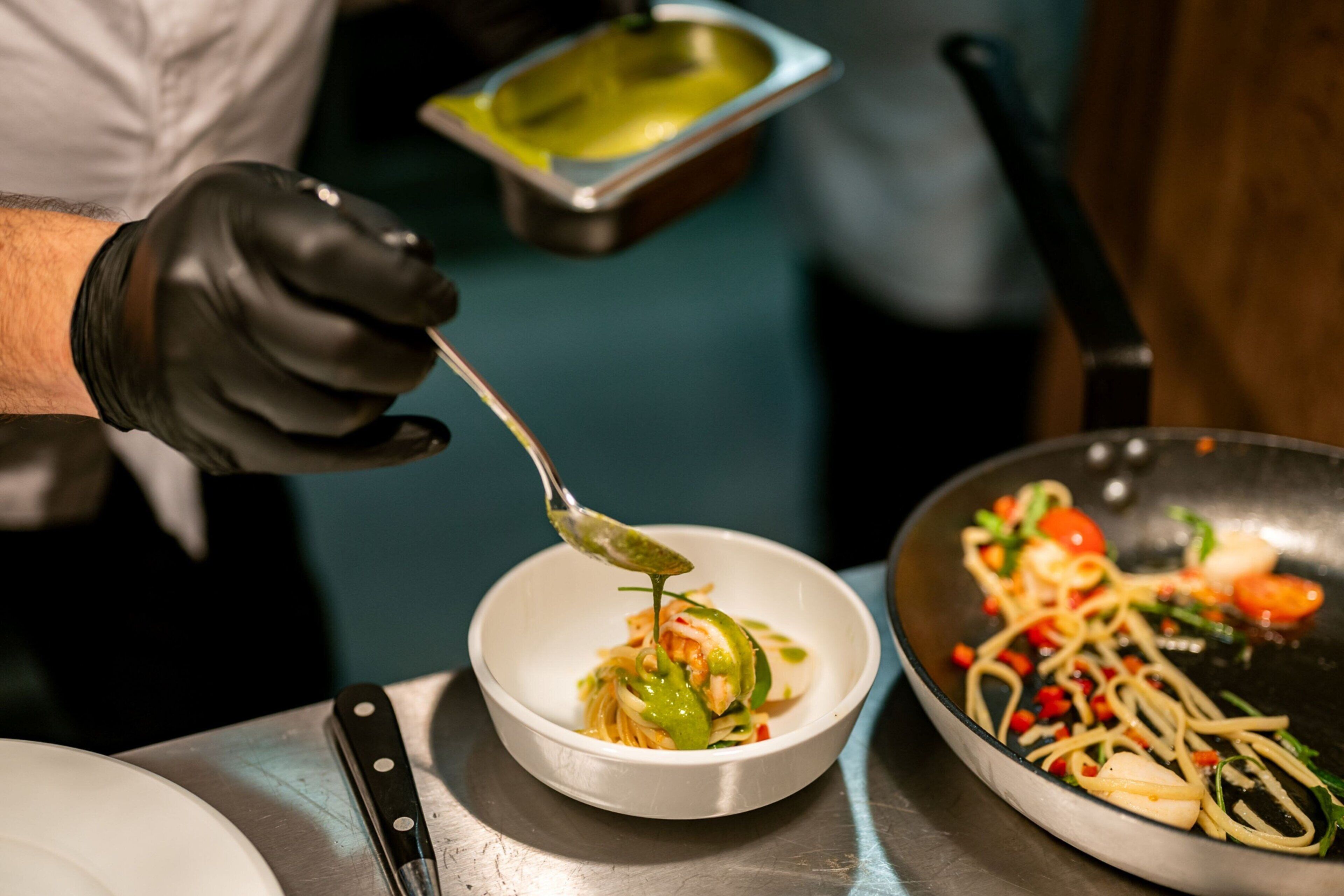 A chef preparing a dish with a spoon, a bowl, and a pan on a stainless steel table.