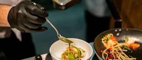 A chef preparing a dish with a spoon, a bowl, and a pan on a stainless steel table.