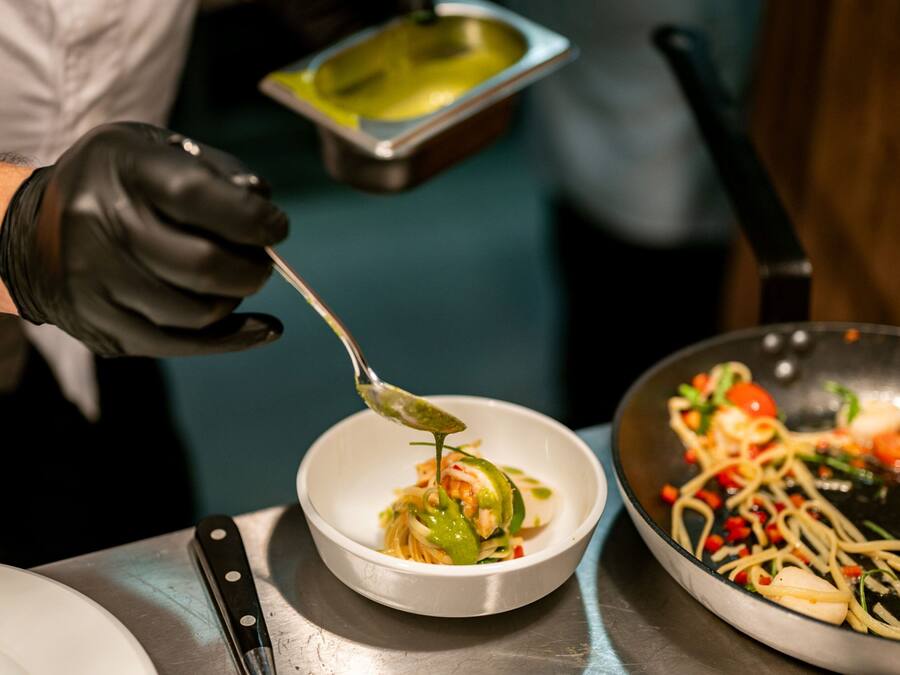 A chef preparing a dish with a spoon, a bowl, and a pan on a stainless steel table.