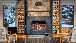A cozy fireplace with logs, a stone wall, and a view of a snowy landscape. There are two wooden chairs and a small table in front of the fireplace. - Four Points by Sheraton Bansko (Bansko)